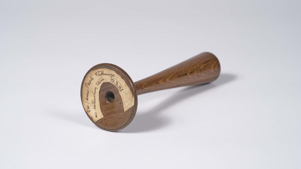 Wooden funnel shaped instrument with a wide base, lying on its side. A hand written label on the base reads 'Paget's Stethoscope'