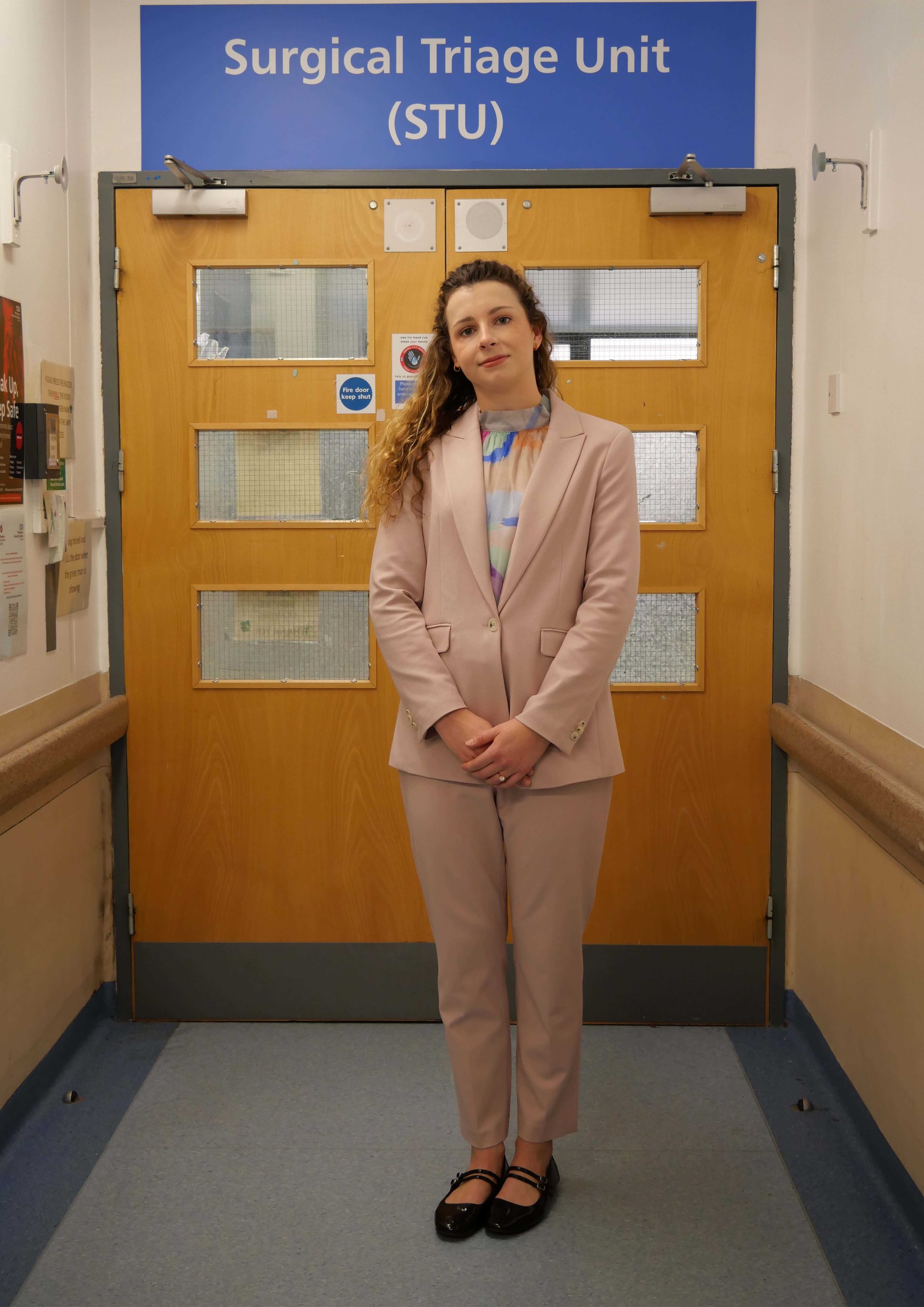 Individual in a pink suit, standing in a hospital corridor in front of a sign saying 'Surgical Triage Unit'