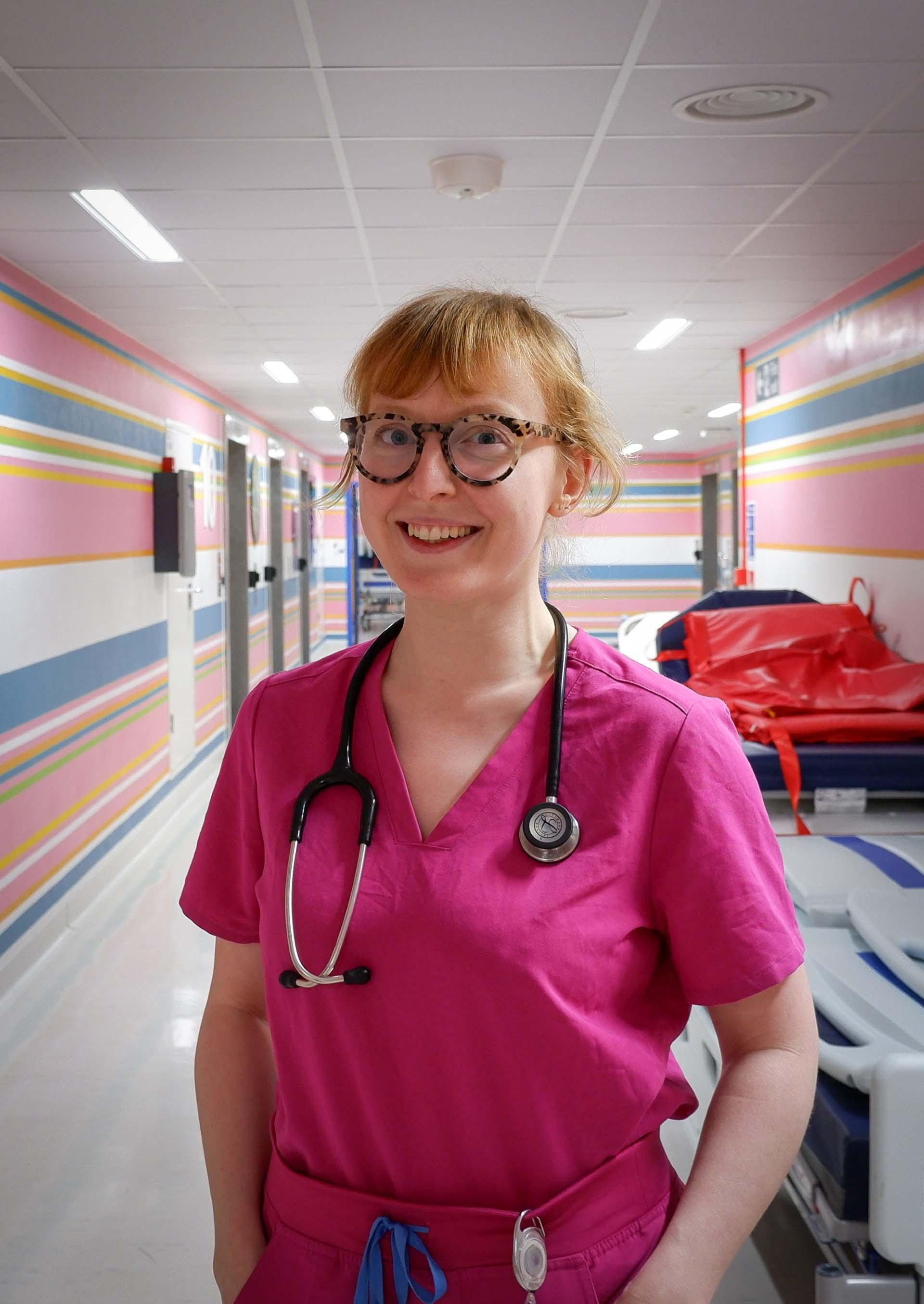 Individual in pink scrubs, wearing a stethoscope, standing in hospital corridor