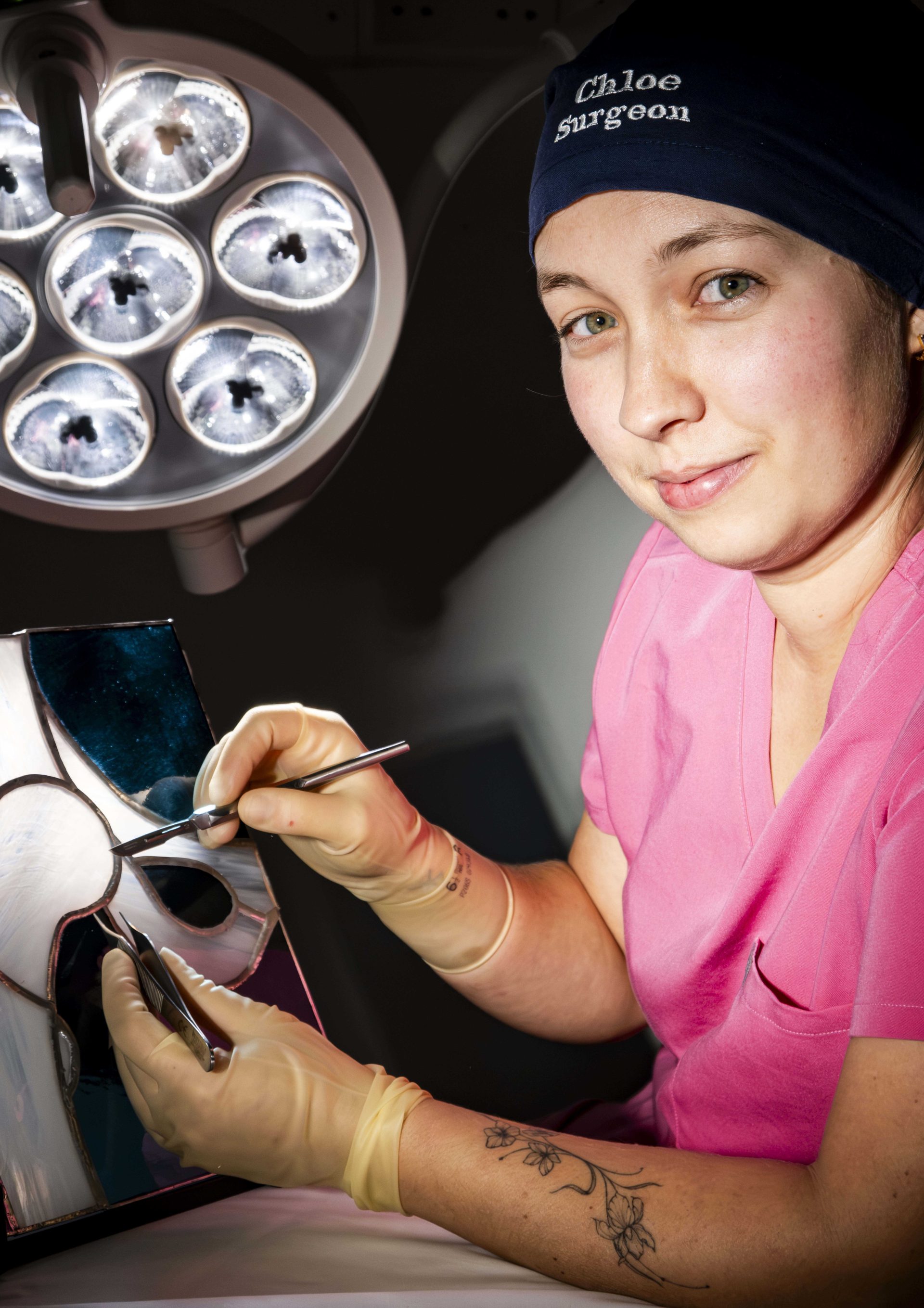 Individual in pink scrubs, sitting in front of a surgical light, with a scalpel and a stain glass depiction of a hip joint