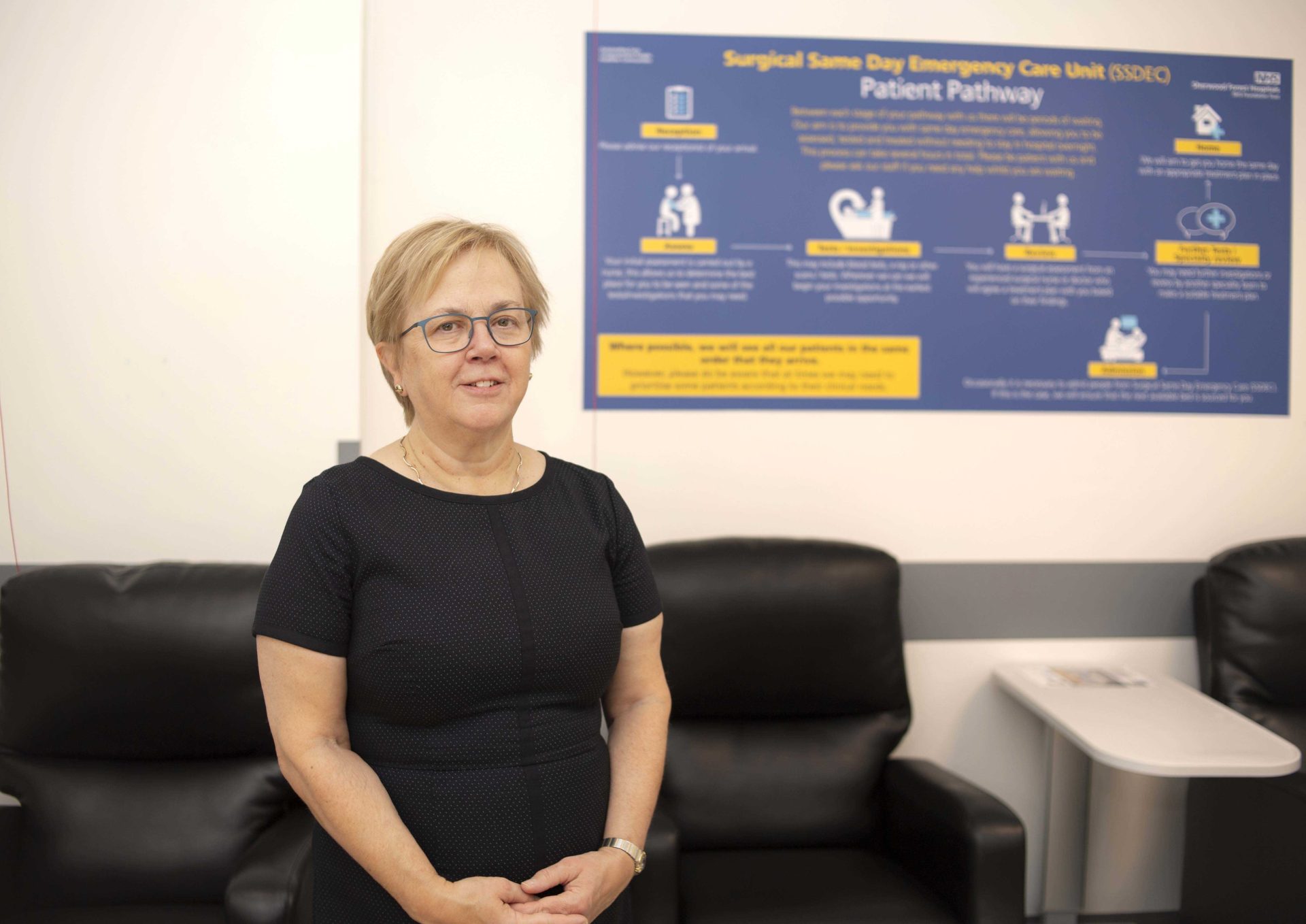 Individual sitting on a black sofa, wearing a black top, in front of a poster describing a 'Surgical Same Day Emergency Care Unit Patient Pathway'
