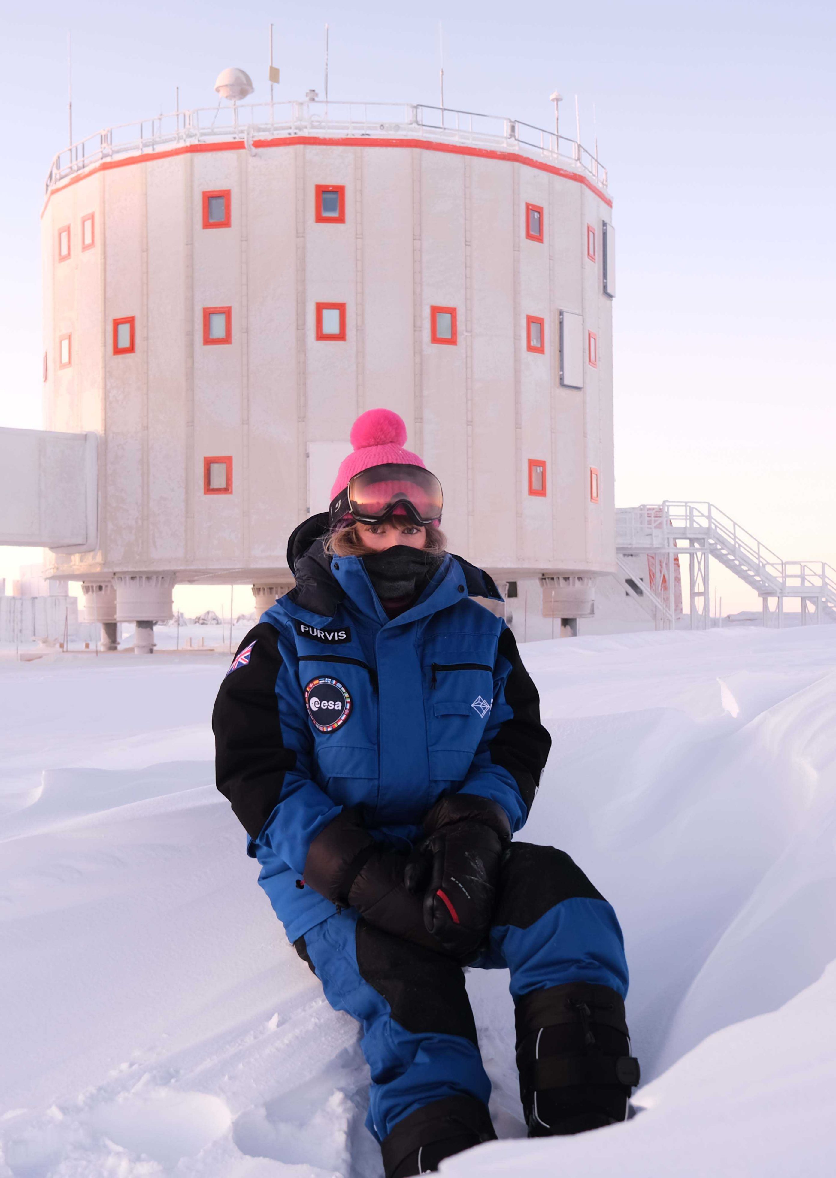 Individual sitting on a snowbank with a pink hat, in front of a large round building