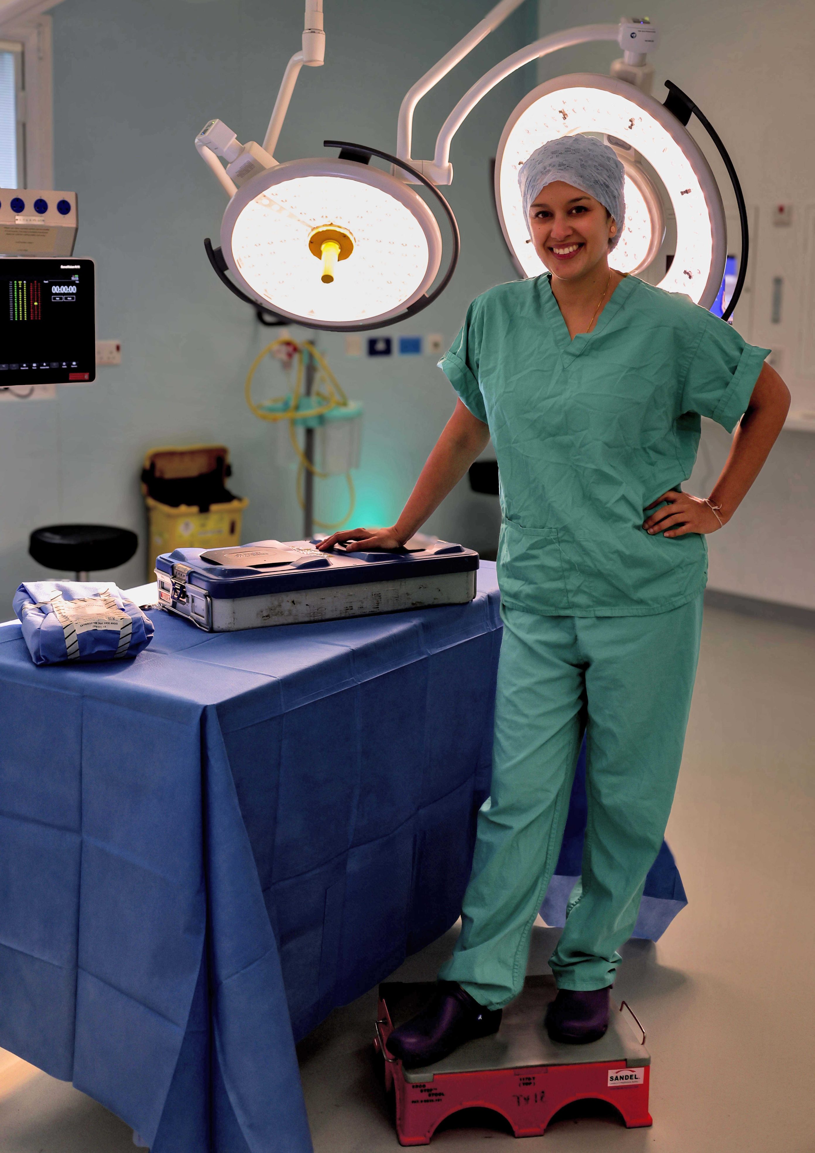 Individual in green scrubs, standing in a surgical theatre next to a draped table, and standing on a low step