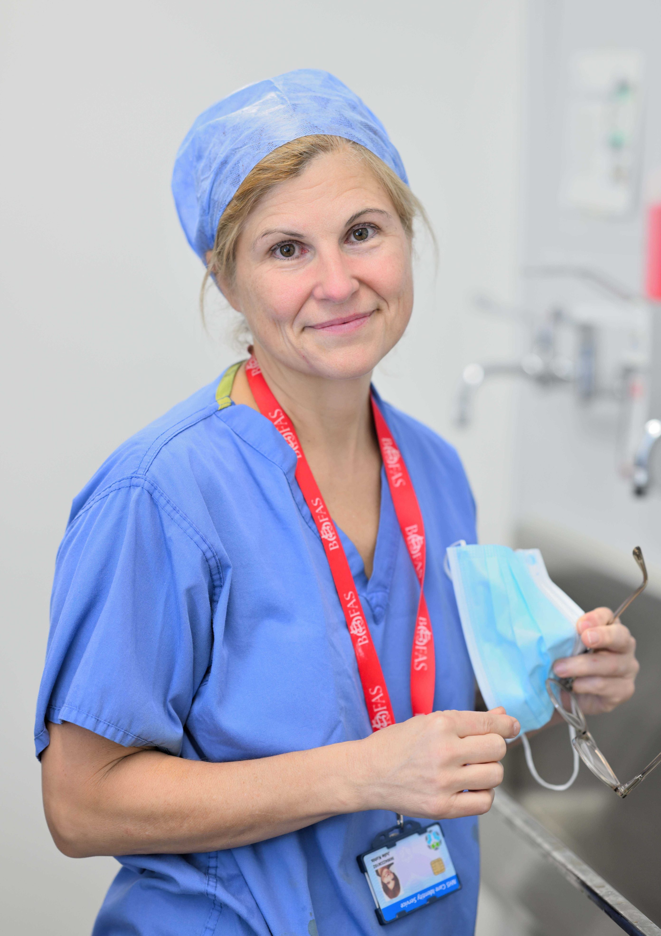 Individual wearing blue scrubs and a lanyard standing in a scrub room in front of a sink