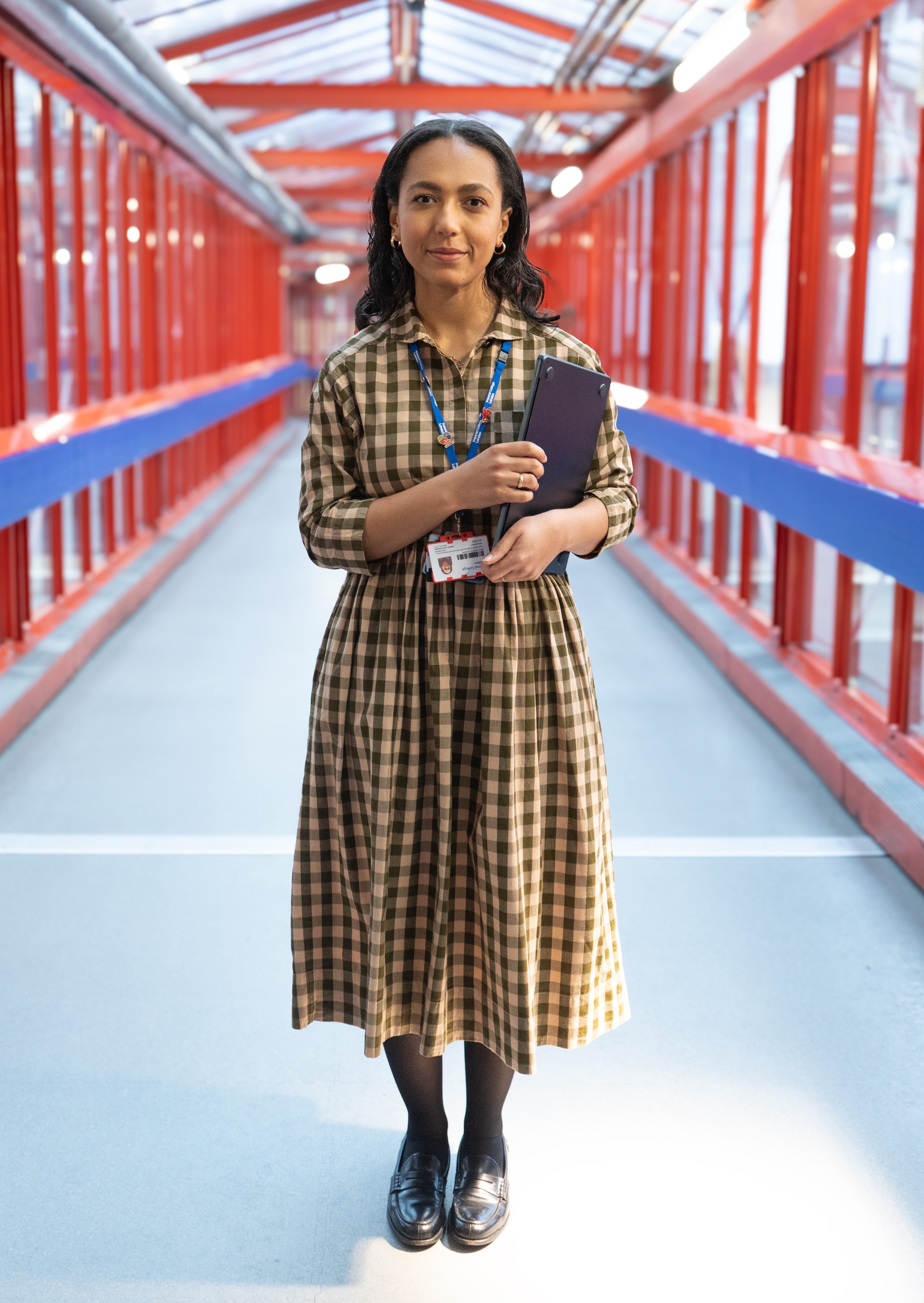 Individual in a brown check dress, standing in a glass lined corridor holding a folder