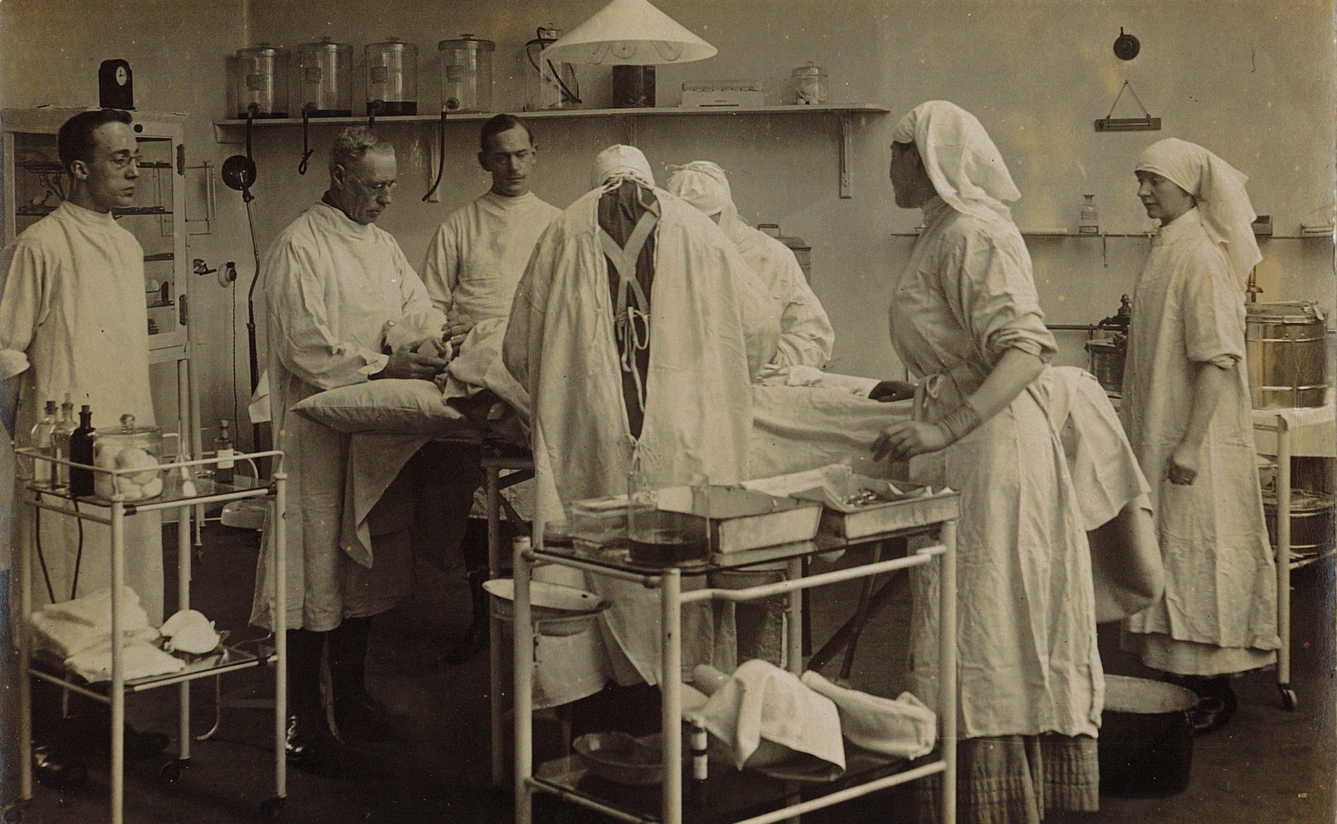 A black and white image of a operating theatre from the 1900s. Six men and two women stand around an operating table dressed in white. They are surrounded by glass bottles and metal equipment,