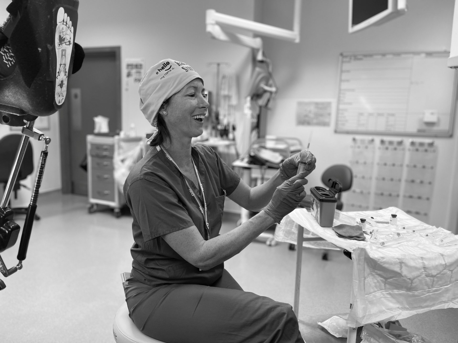 A black and white image of a white woman sat on a stool in an operating room. She is wearing surgical scrubs and cap and smiles as she uses a tool in her hand.